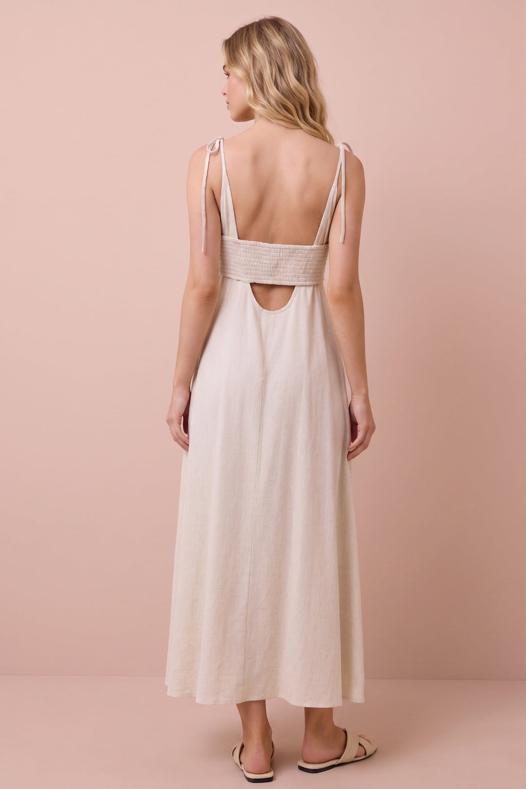 A woman shown from the back wearing a light beige maxi dress with thin shoulder straps, a wide textured band across the upper back, and a cutout at the center of the back, standing against a solid pale pink studio backdrop.