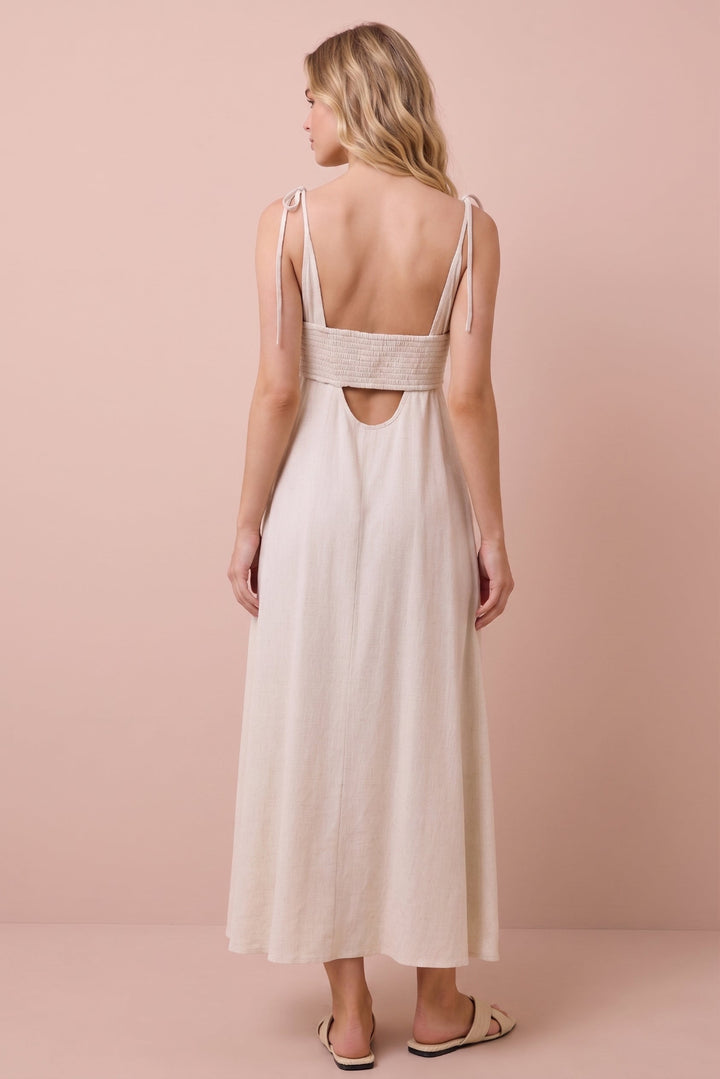 A woman shown from the back wearing a light beige maxi dress with thin shoulder straps, a wide textured band across the upper back, and a cutout at the center of the back, standing against a solid pale pink studio backdrop.