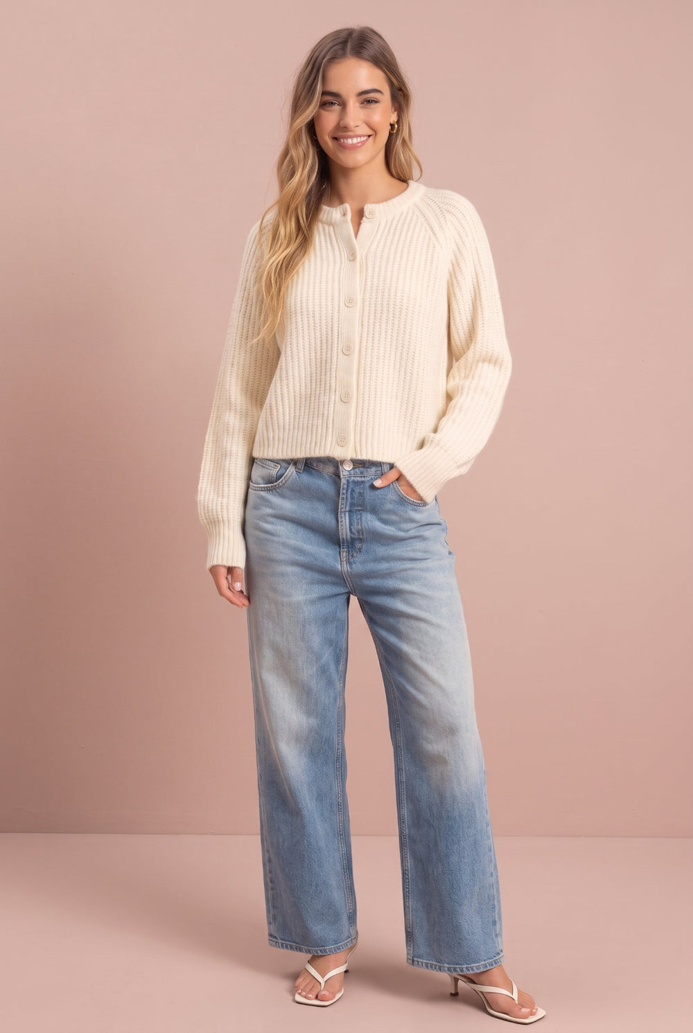 Woman wearing cream ribbed cardigan and light blue jeans against pink background, front view.