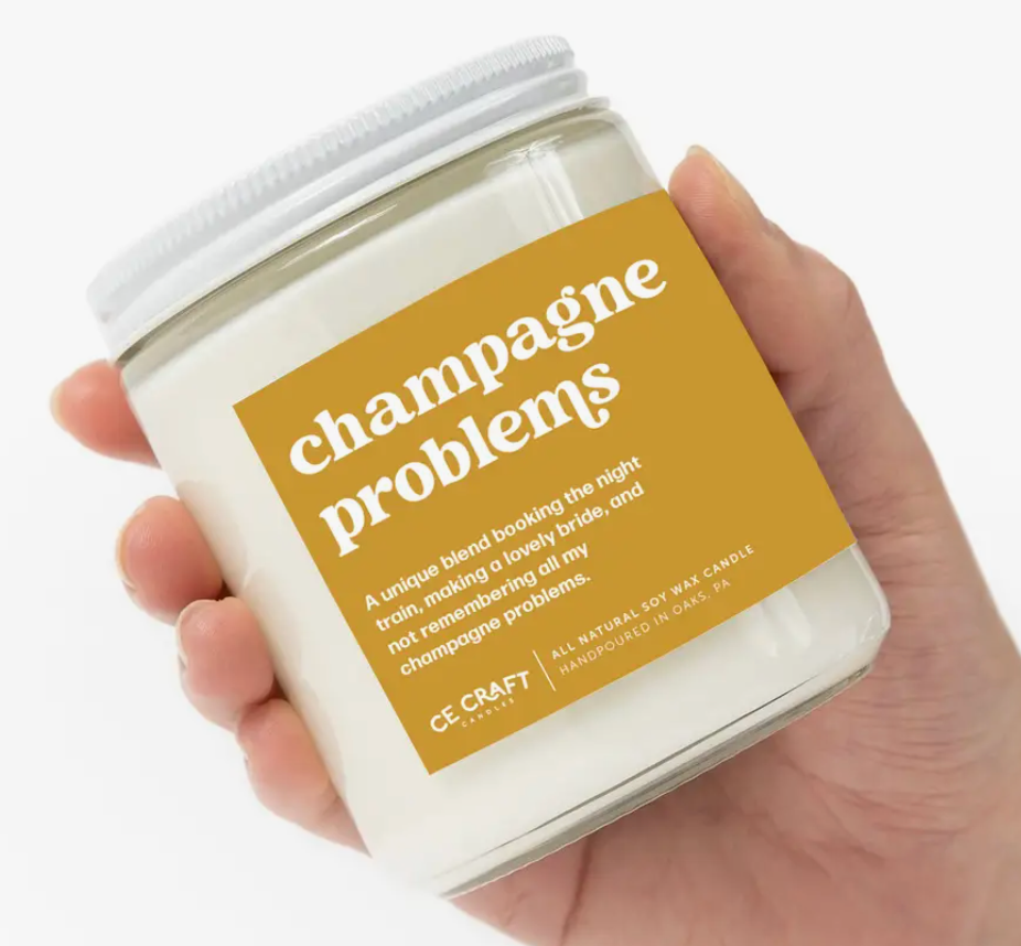 A candle jar with a yellow and white label that says "Champagne problems", featuring a unique blend of scents.