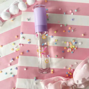 A lilac-colored wand-style lip gloss applicator filled with a clear, sparkling liquid, set against a pink and white striped background with colorful confetti-like shapes.