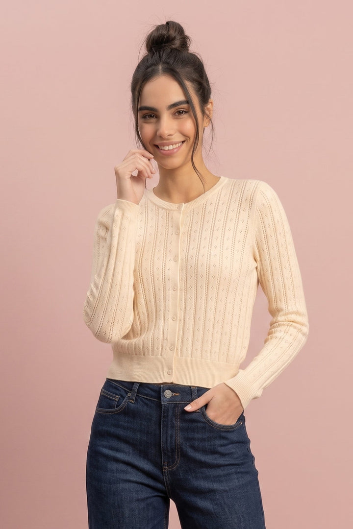 A woman modeling a cream ribbed knit cardigan with button-front closure and long sleeves, paired with dark blue jeans, against a solid dusty pink studio background.