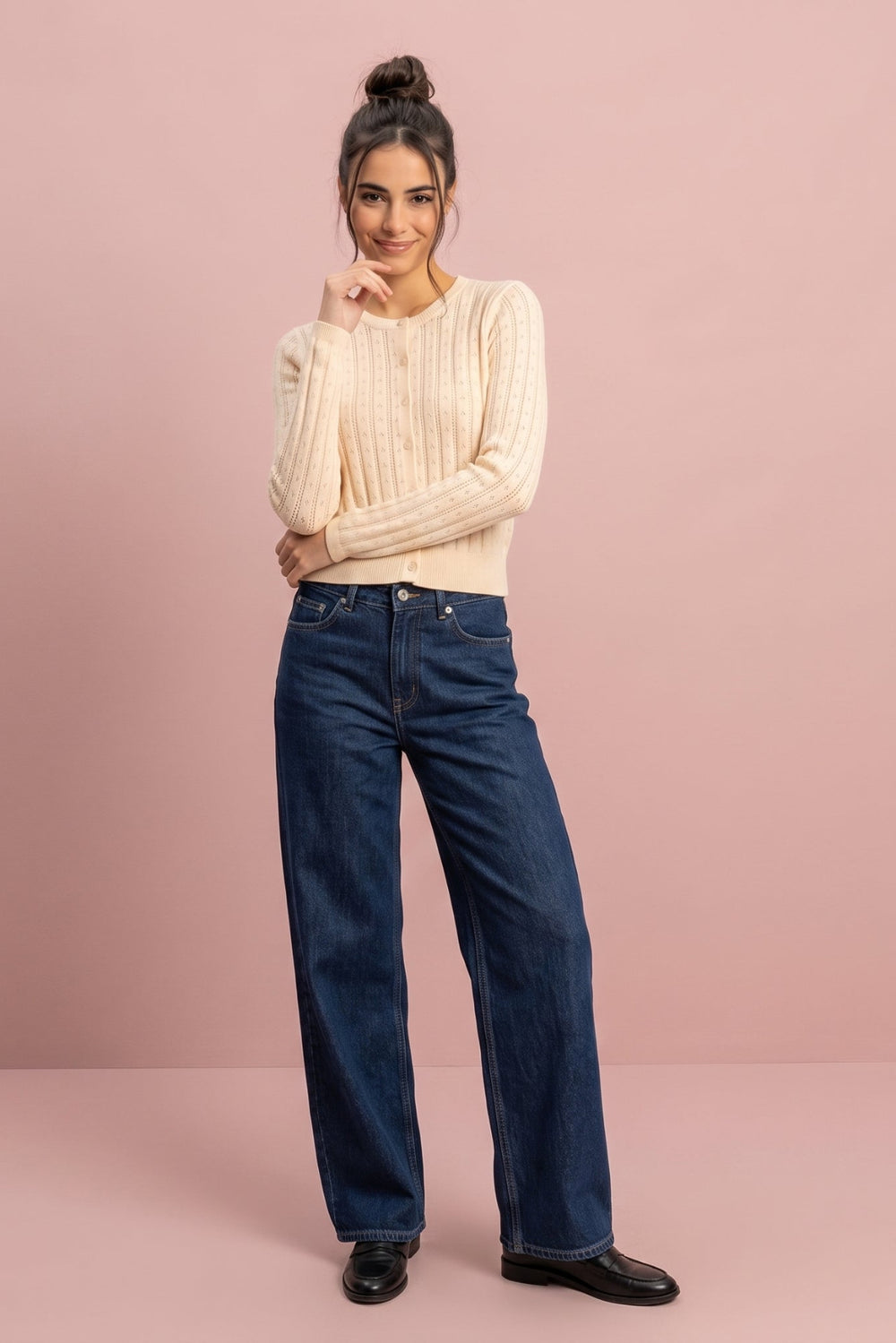 A woman stands against a solid dusty pink studio background wearing a cream ribbed knit sweater, dark blue wide-leg jeans, and black leather shoes.