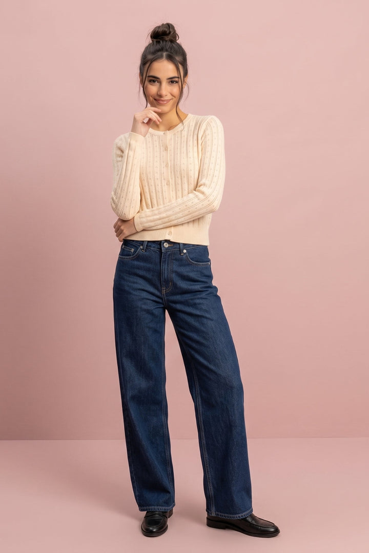 A woman stands against a solid dusty pink studio background wearing a cream ribbed knit sweater, dark blue wide-leg jeans, and black leather shoes.