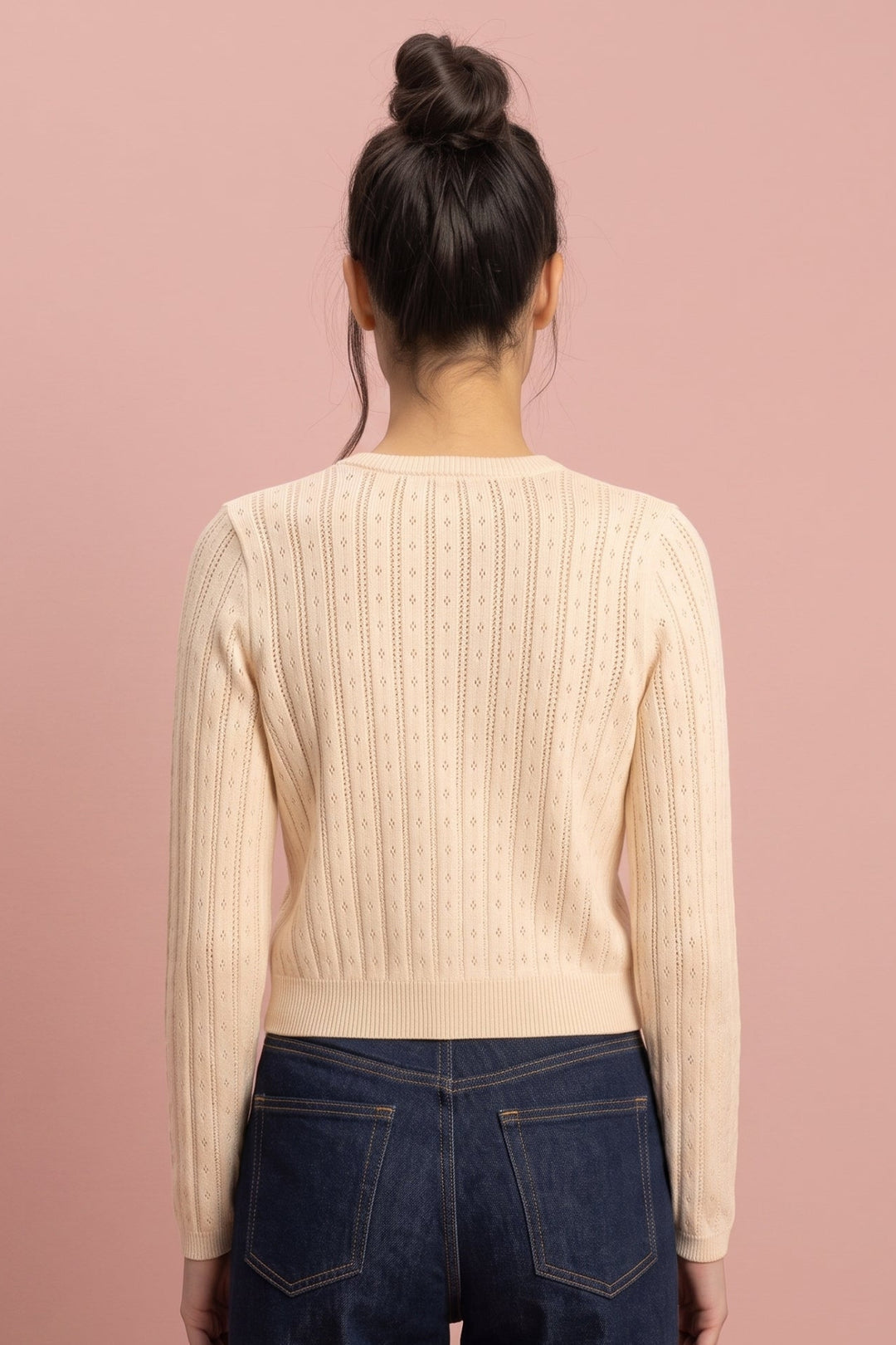 A woman viewed from behind wearing a cream ribbed knit sweater and dark blue jeans, standing against a solid dusty pink background.