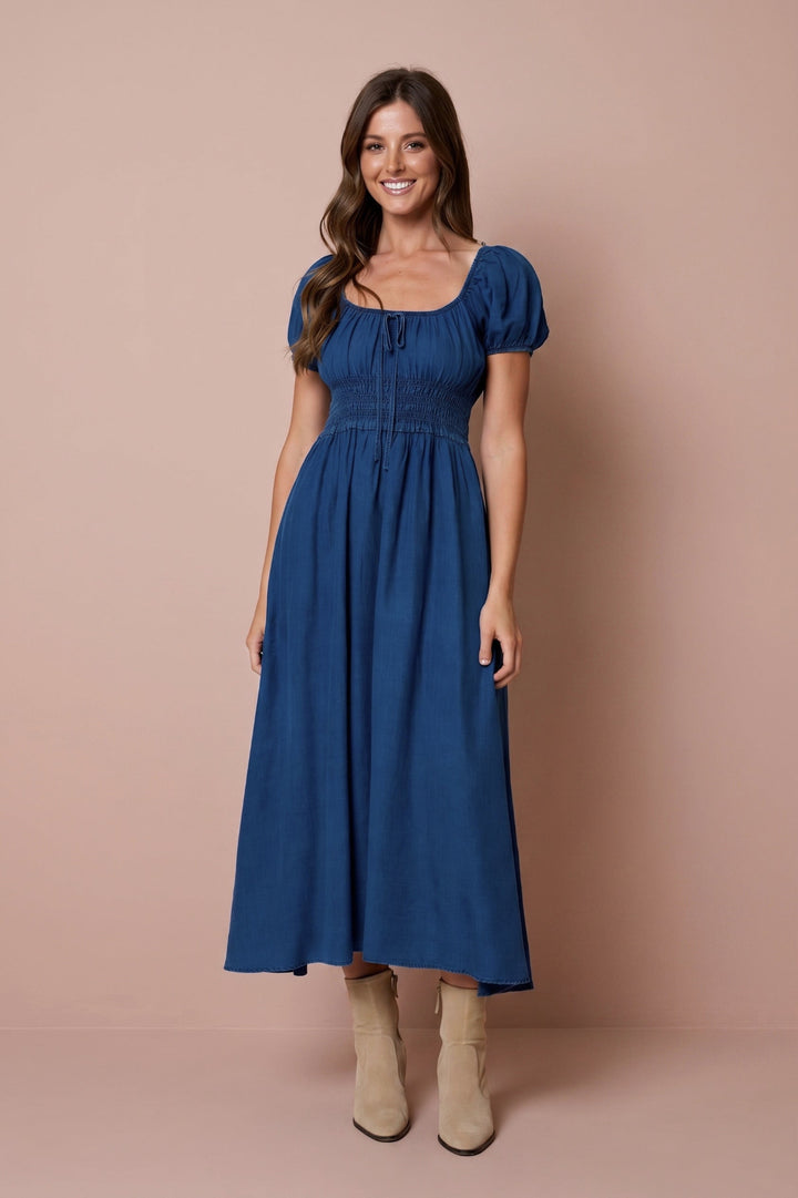 A woman modeling a blue midi dress with puff sleeves, a smocked waist, and a square neckline, paired with beige ankle boots, set against a solid dusty pink studio backdrop.