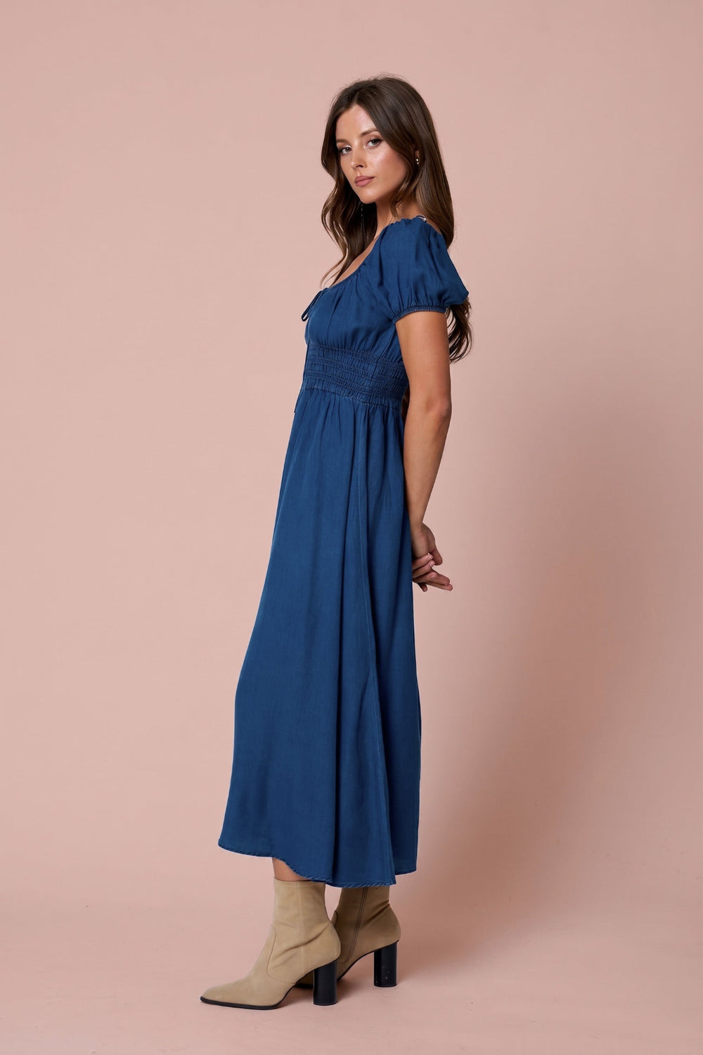 A woman modeling a blue midi dress with short puff sleeves and a gathered waist, paired with beige ankle boots, against a solid dusty pink studio background.