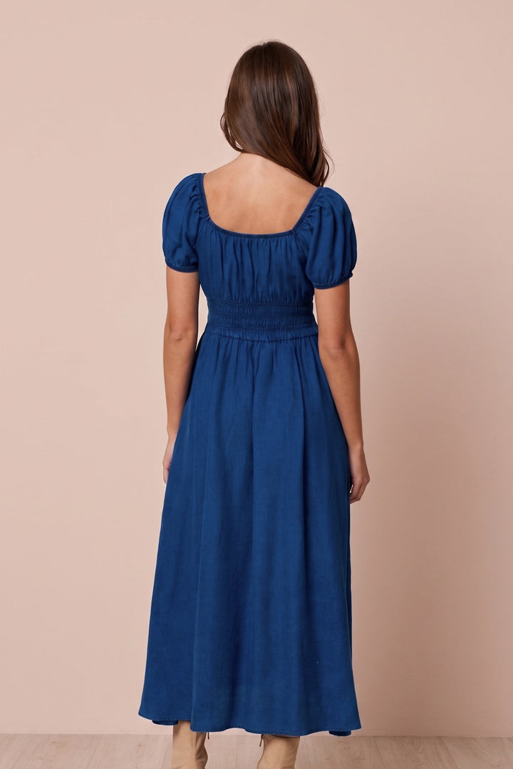 A woman viewed from the back wearing a royal blue midi dress with puff sleeves, a square neckline, and a smocked waist, paired with beige ankle boots, set against a solid beige studio background.