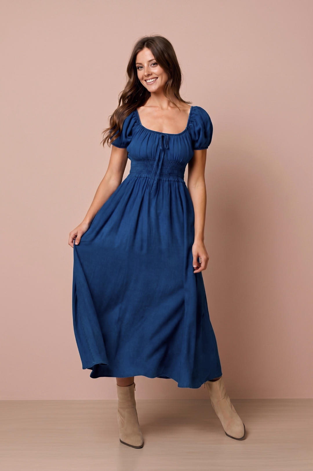A woman modeling a midi-length blue dress with puff sleeves, a square neckline, and a gathered waist, paired with beige ankle boots against a solid dusty pink studio backdrop.