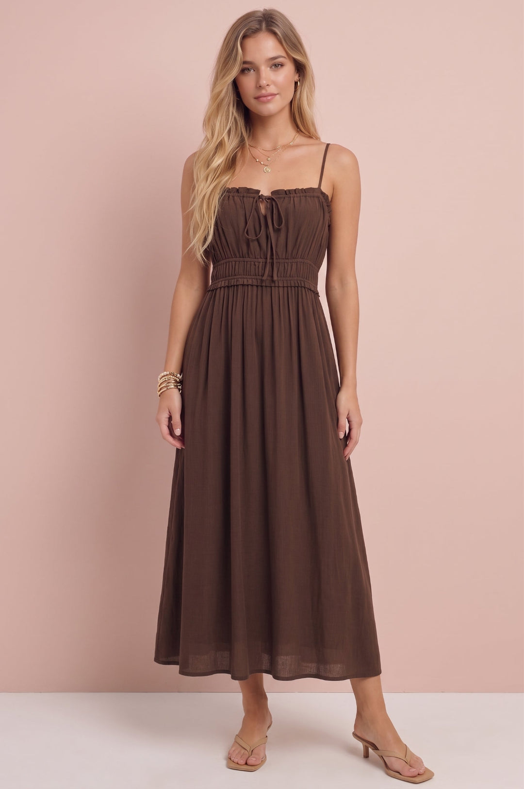 A woman modeling a brown midi dress with thin spaghetti straps, a smocked bodice, and a flowing skirt, standing against a solid pale pink studio backdrop.