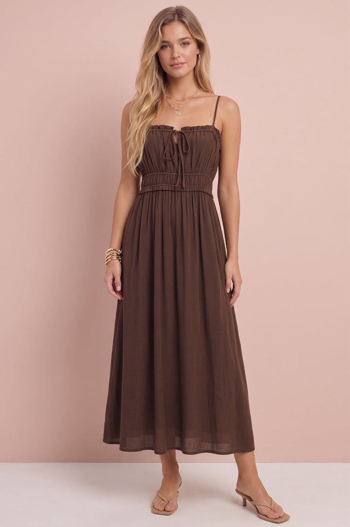 A woman modeling a brown midi dress with thin spaghetti straps, a smocked bodice, and a flowing skirt, standing against a solid pale pink studio backdrop.