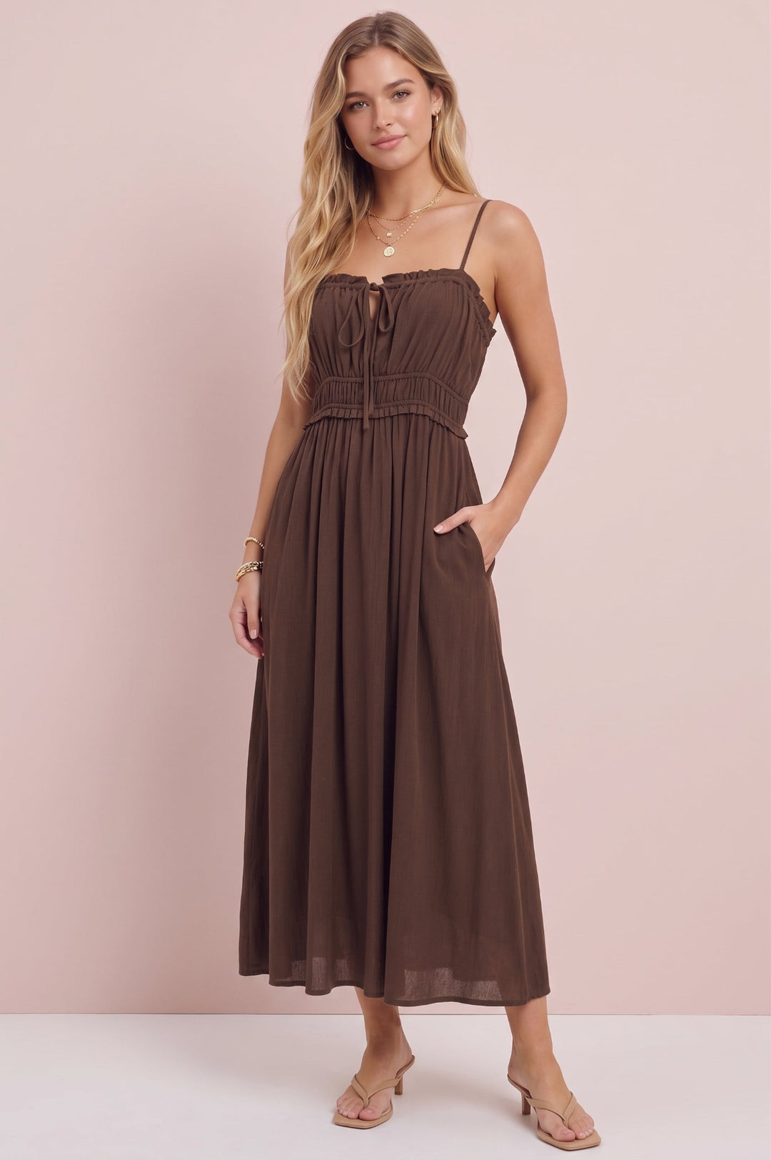 A woman modeling a brown midi dress with thin straps, a smocked bodice, and side pockets, standing against a solid pale pink background.