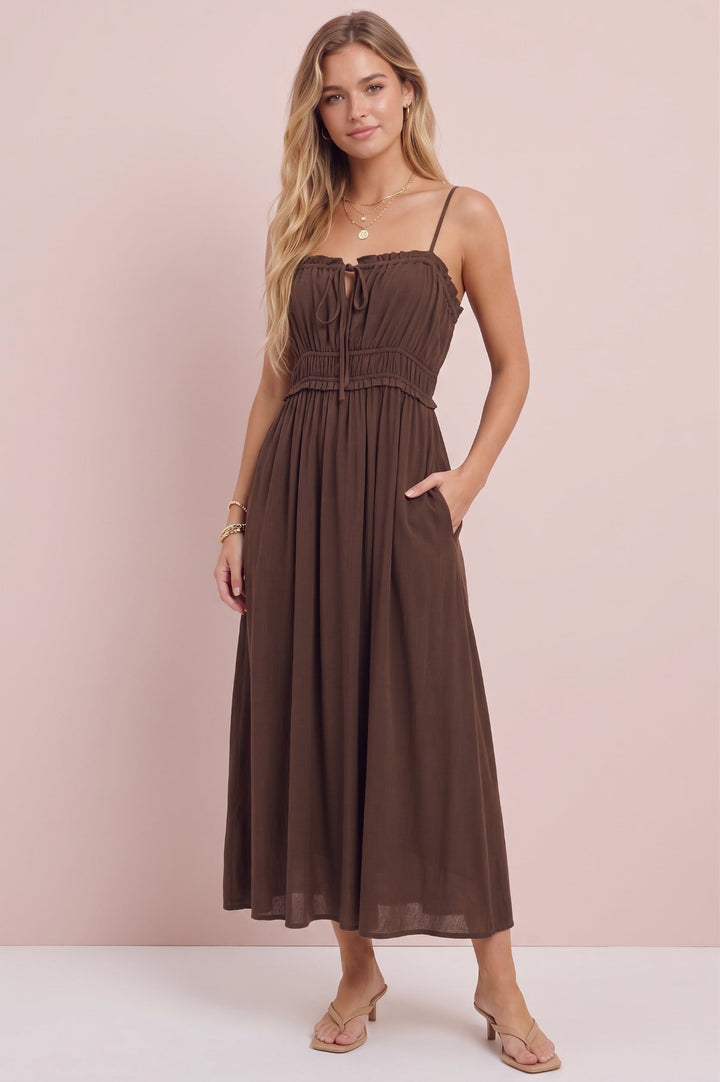 A woman modeling a brown midi dress with thin straps, a smocked bodice, and side pockets, standing against a solid pale pink background.