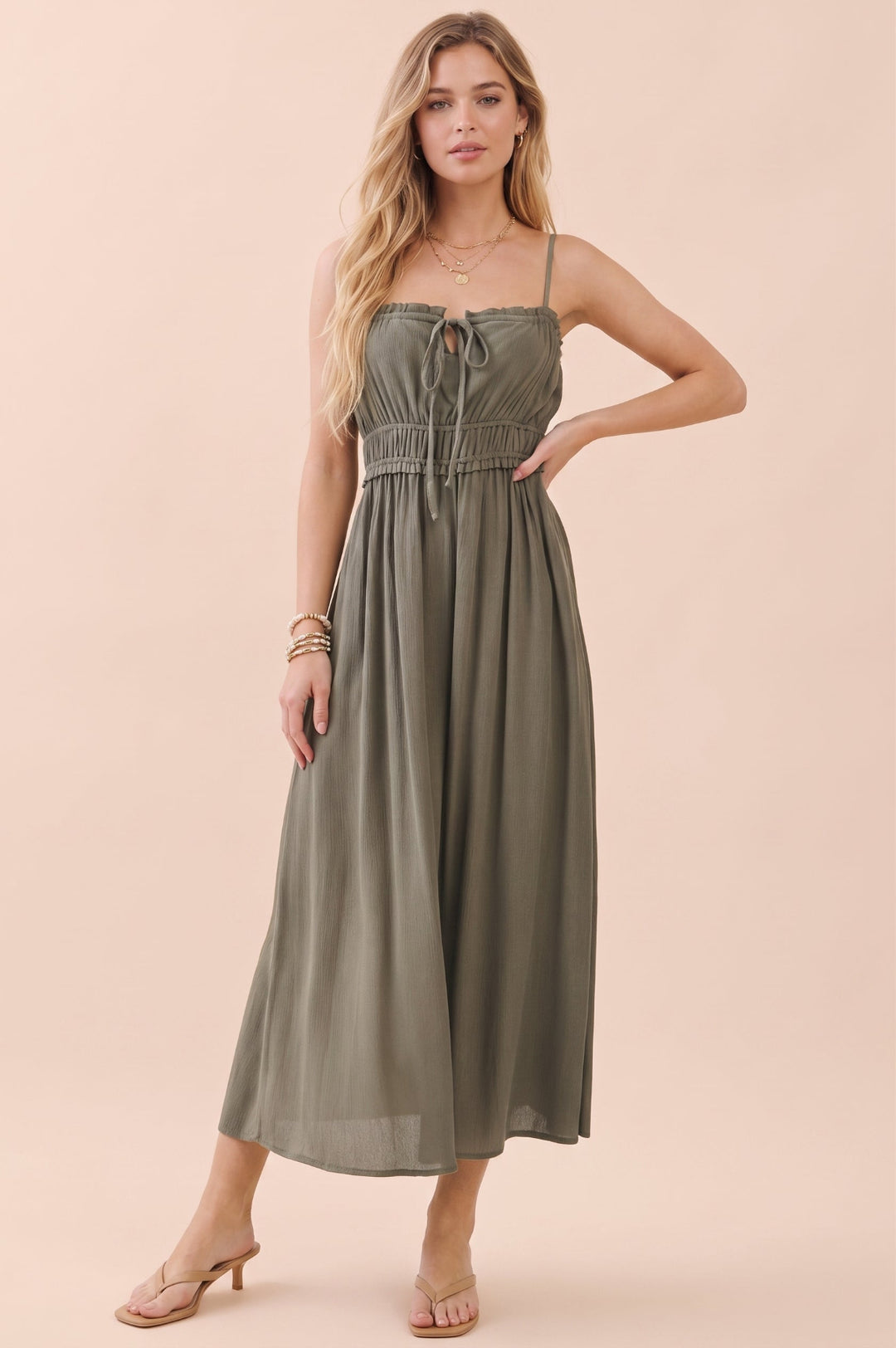 A woman modeling a sleeveless olive green midi dress with thin straps, a tie-front bodice, and a gathered waist, posed against a solid pale peach background.
