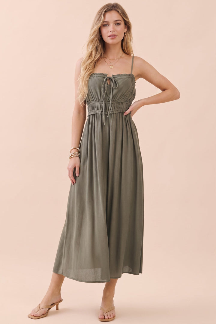 A woman modeling a sleeveless olive green midi dress with thin straps, a tie-front bodice, and a gathered waist, posed against a solid pale peach background.
