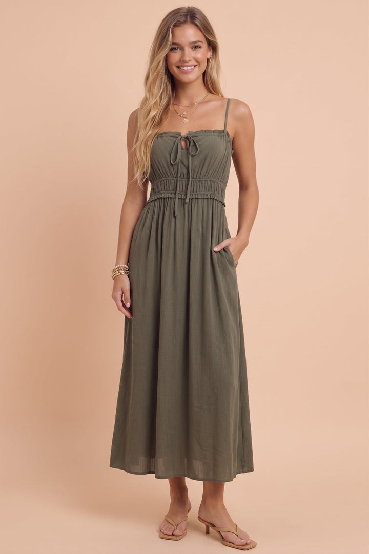 A woman modeling an olive green midi dress with spaghetti straps, a tie-front detail, a gathered waist, and side pockets, set against a solid peach studio background.