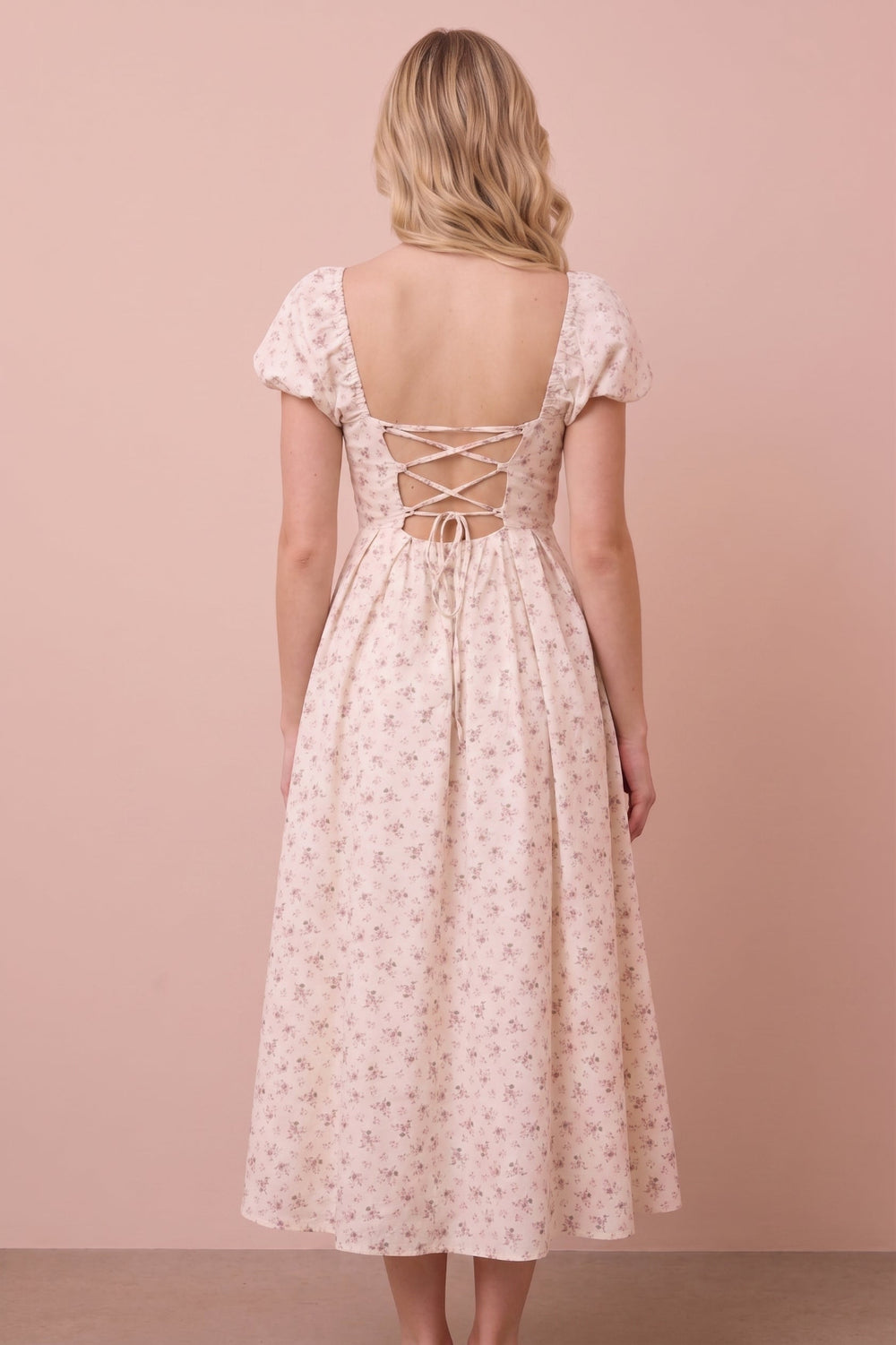 A woman viewed from the back wearing a light blush pink midi dress with a small floral print and lace-up open back, standing against a solid dusty pink background.