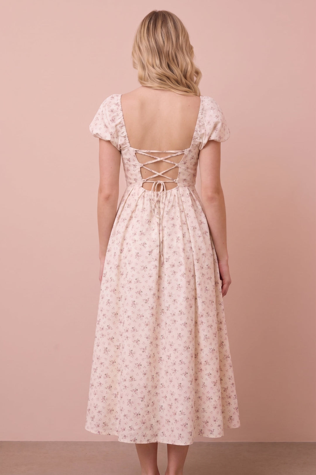 A woman viewed from the back wearing a light blush pink midi dress with a small floral print and lace-up open back, standing against a solid dusty pink background.