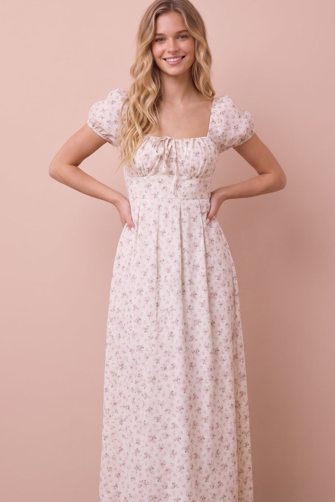 A woman modeling a long, cream-colored maxi dress with a delicate pink floral pattern, puff sleeves, and a square neckline, set against a solid dusty pink background.