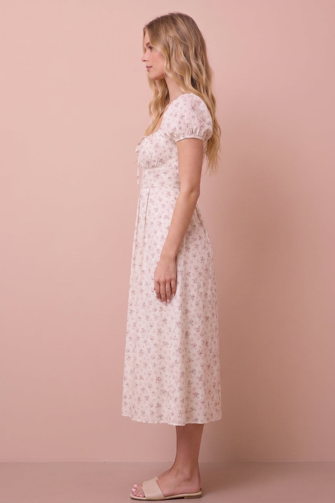 A woman stands in profile wearing a short-sleeve midi dress with a small floral print. The dress is off-white with a delicate pink and green floral pattern.