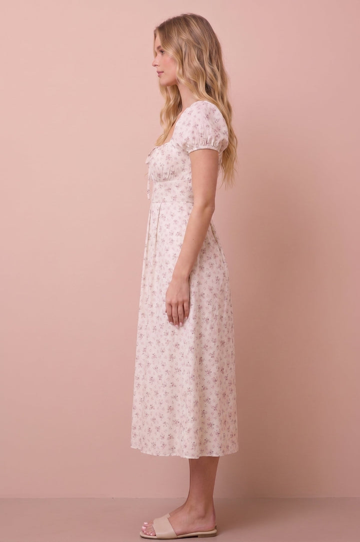 A woman stands in profile wearing a short-sleeve midi dress with a small floral print. The dress is off-white with a delicate pink and green floral pattern.