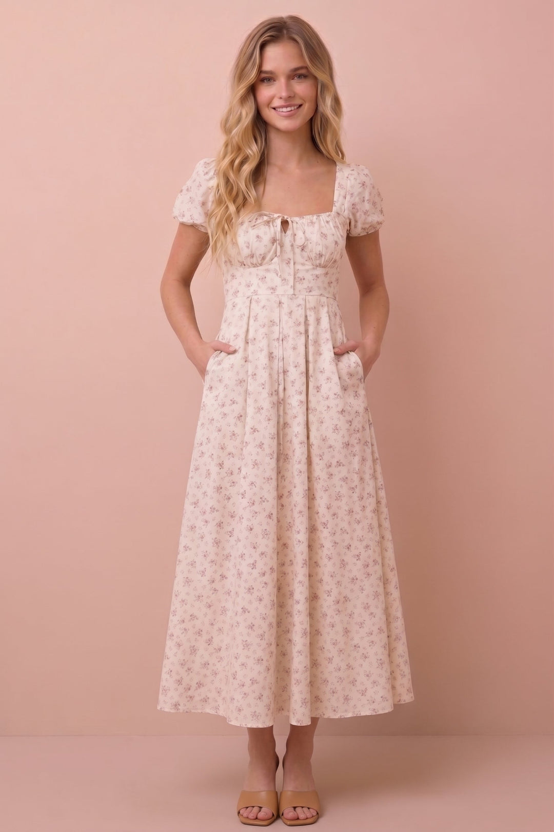 A woman modeling a short-sleeve midi dress with a small floral print in beige and pink, standing against a solid pink studio backdrop.