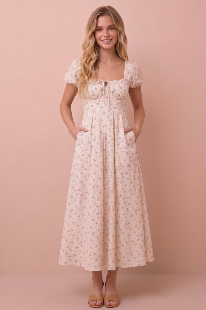 A woman modeling a short-sleeve midi dress with a small floral print in beige and pink, standing against a solid pink studio backdrop.