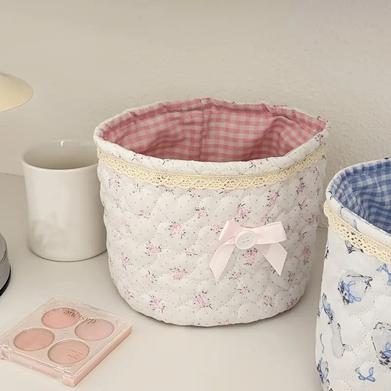 A white storage basket with a floral pattern and lace detailing, featuring a pink and white gingham lining and a small bow accent.
