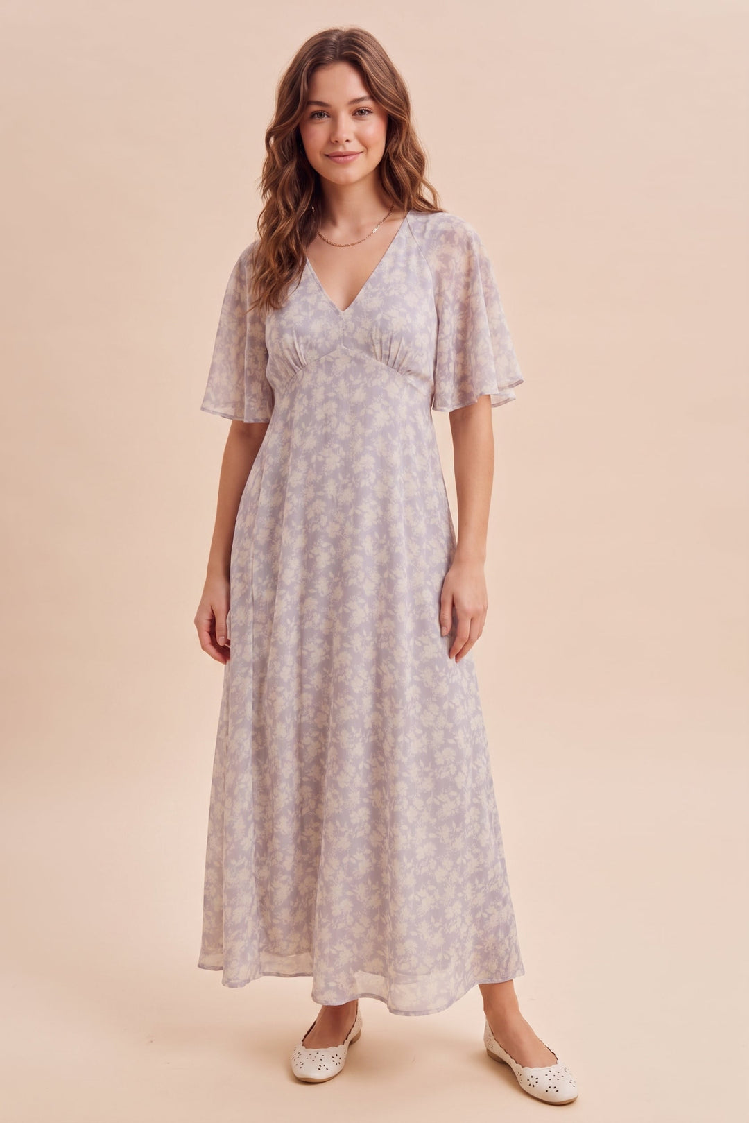 A woman modeling a light lavender-gray maxi dress with a subtle floral pattern, short flutter sleeves, and a V-neckline, standing against a solid beige background.