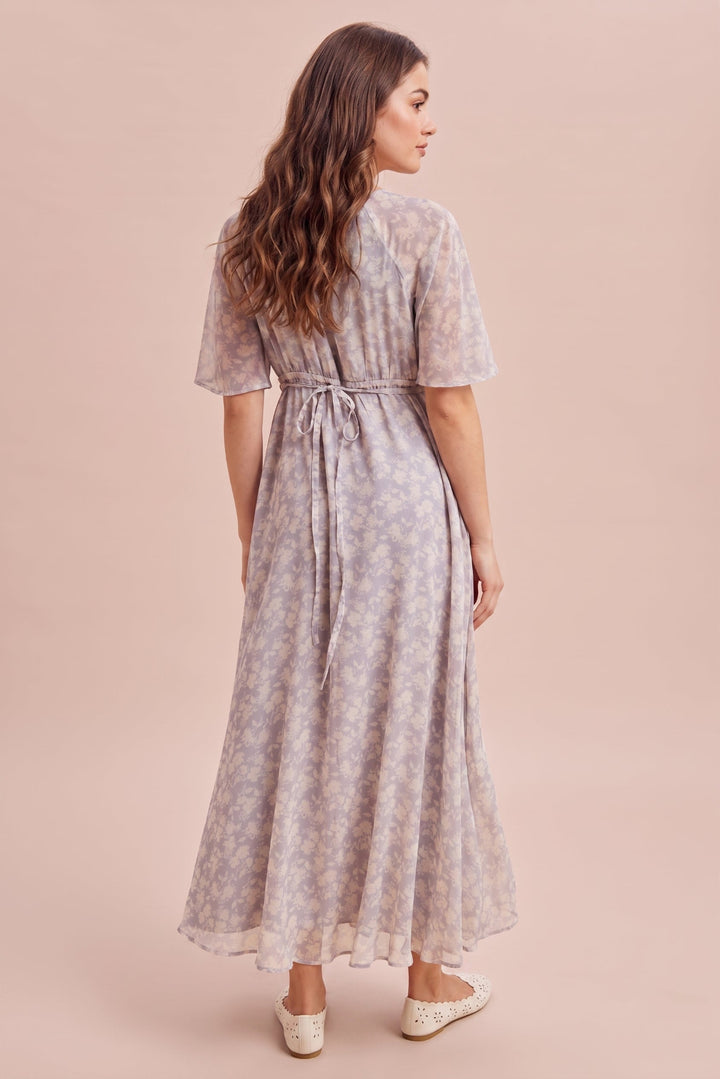A woman wearing a light lavender floral print midi dress with short sleeves and a tie waist, shown from the back against a solid pale pink background.