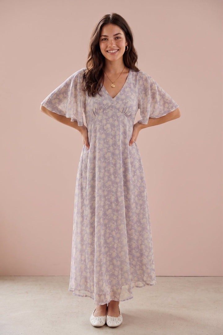 A woman modeling a light lavender maxi dress with a delicate floral pattern, featuring flutter sleeves and a V-neckline, set against a solid dusty pink studio background.