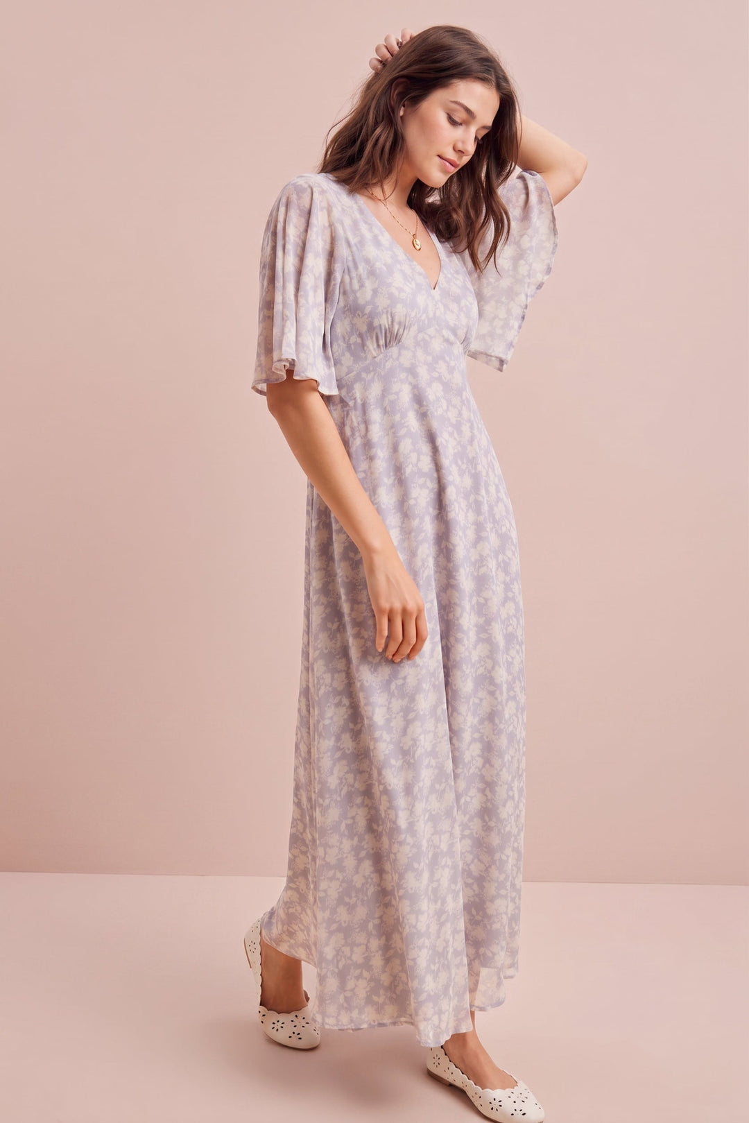 A woman modeling a light lavender and white floral print maxi dress with short puff sleeves and a V-neckline, standing against a solid pale pink studio backdrop.