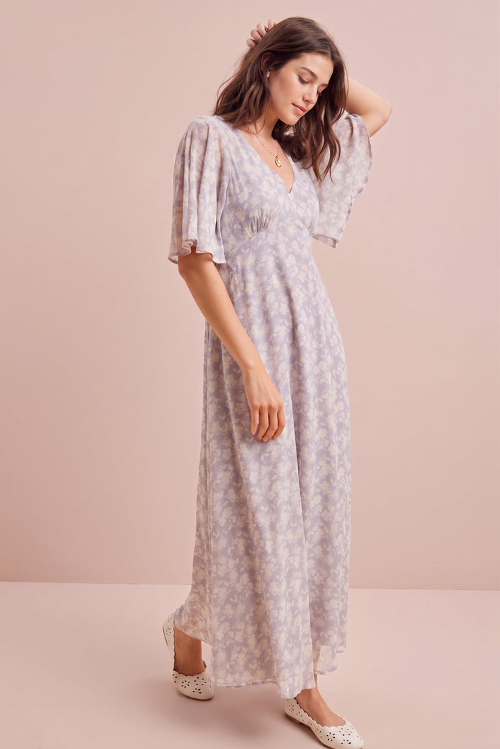 A woman modeling a light lavender and white floral print maxi dress with short puff sleeves and a V-neckline, standing against a solid pale pink studio backdrop.