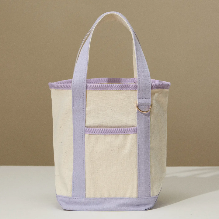 A lightweight, eco-friendly canvas tote in a neutral beige color with lavender trim, featuring a simple design with a metal ring detail.