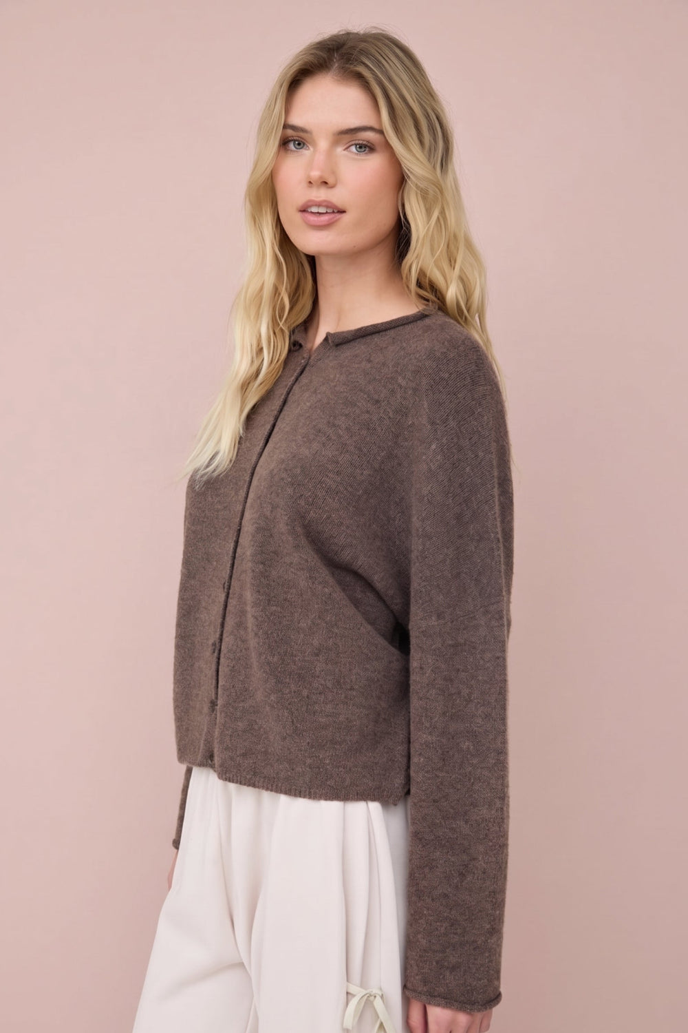 A woman modeling a cropped, long-sleeve knit cardigan in a heathered brown color, paired with cream-colored pants, set against a solid dusty pink background.