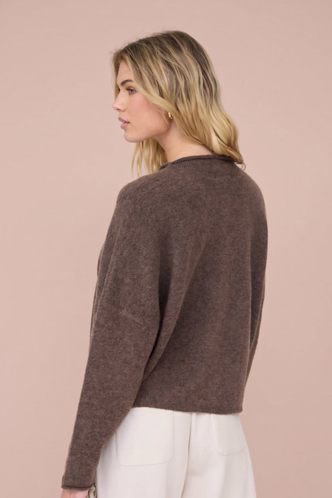 A woman shown from the back wearing a cropped, long-sleeve knit sweater in a warm brown color, paired with white pants, against a solid pale pink background.