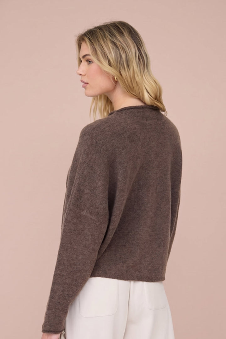 A woman shown from the back wearing a cropped, long-sleeve knit sweater in a warm brown color, paired with white pants, against a solid pale pink background.