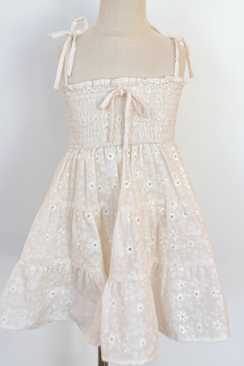 A white sleeveless girls' dress with a smocked bodice, tie shoulder straps, and an eyelet lace skirt with a ruffled hemline.
