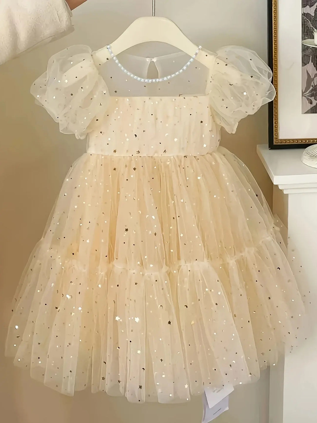 A girls' cream-colored tulle dress with sparkly gold star patterns throughout, featuring puffy short sleeves and a ruffled skirt.