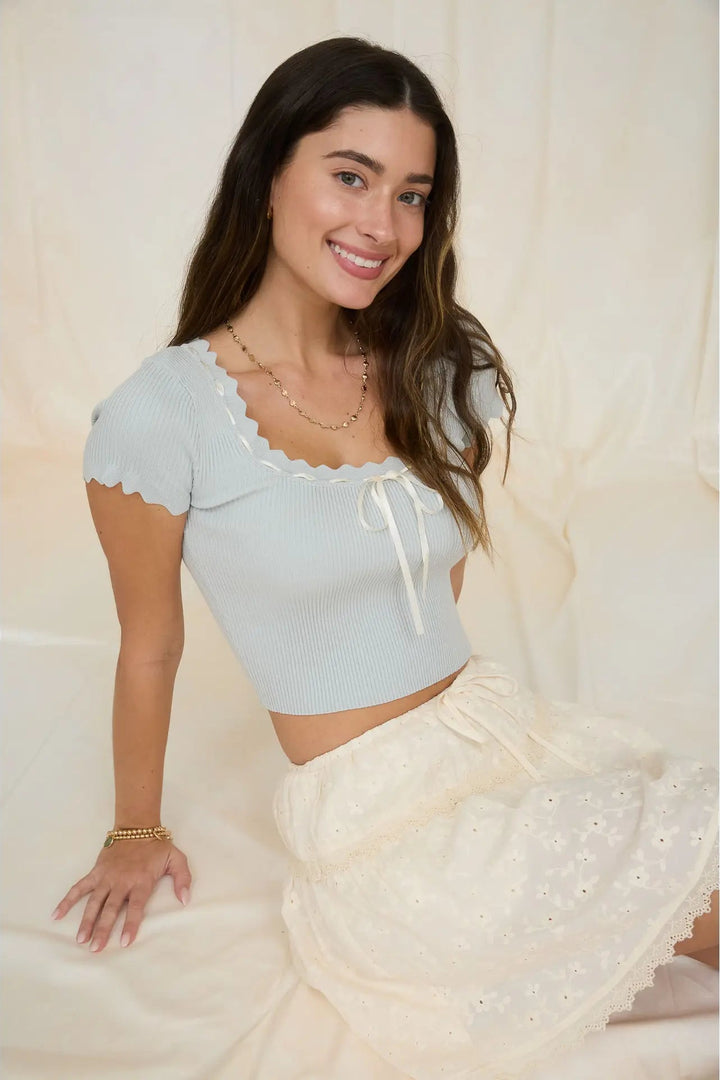 Woman wearing a light blue top and white lace skirt with a plain background