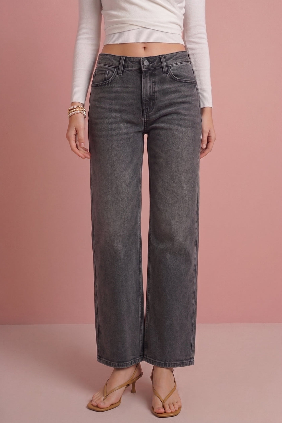 A model wearing dark gray wide-leg cropped jeans, a white long-sleeve crop top, and tan heeled sandals, standing against a solid pink studio background.