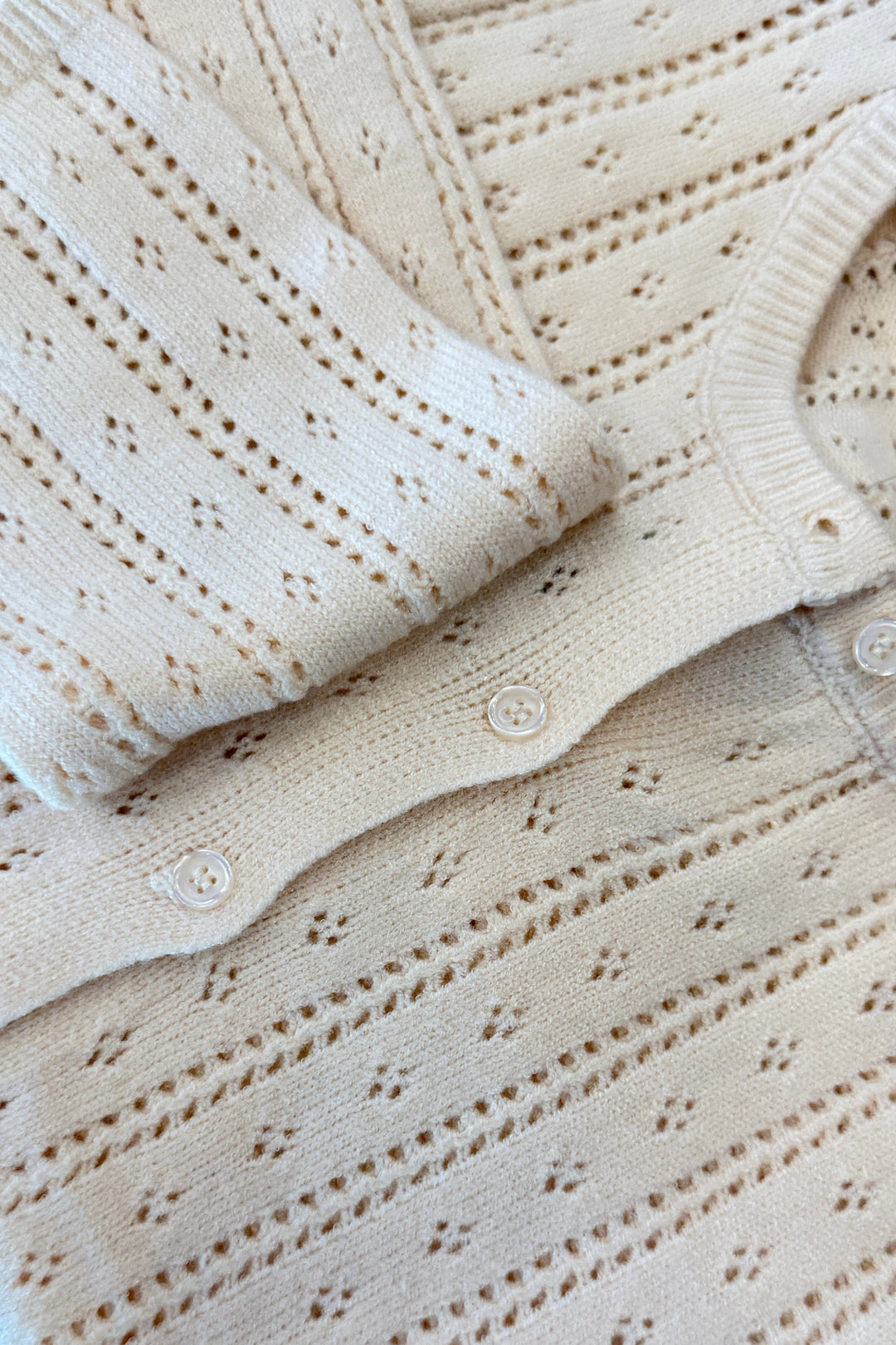 Cream pointelle knit cardigan with button details, folded flat lay view.
