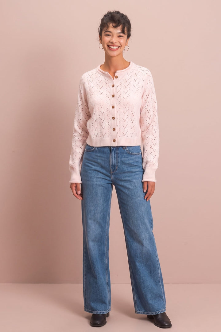 A woman modeling a light pink cable-knit cropped cardigan with brown buttons, paired with blue wide-leg jeans and black shoes, standing against a solid dusty pink studio backdrop.
