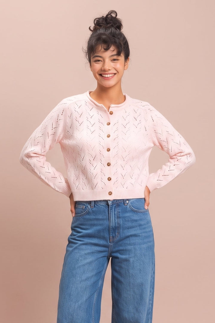 A woman modeling a light pink long-sleeve knit cardigan with a textured chevron pattern and brown buttons, paired with high-waisted blue wide-leg jeans, against a solid dusty pink studio background.