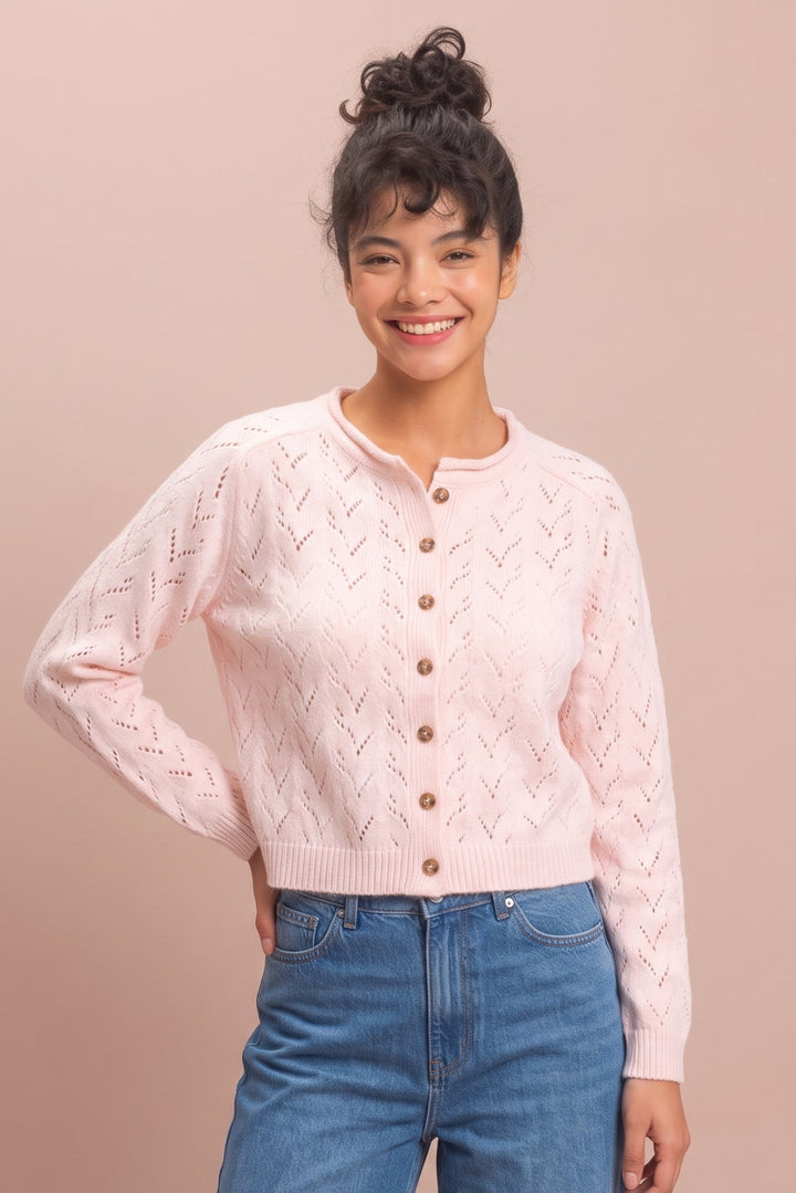 A woman modeling a light pink knit cardigan with brown buttons, paired with blue jeans, against a solid pale pink background.