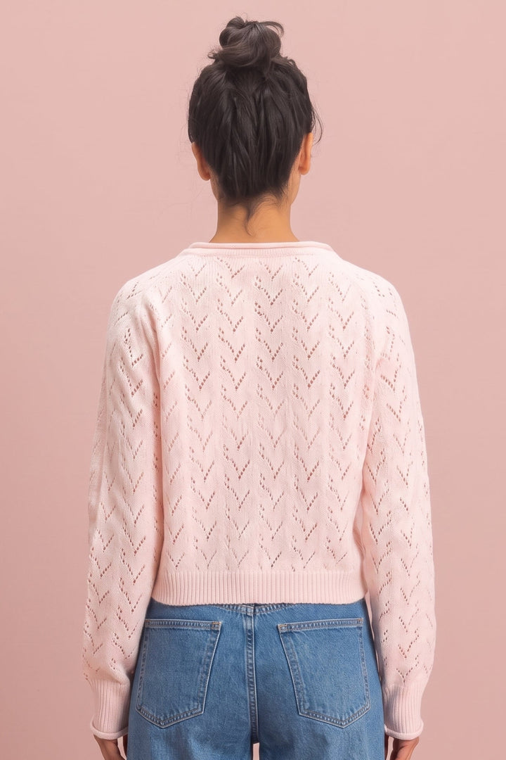 A woman viewed from behind wearing a light pink cable-knit sweater and blue jeans against a solid pink background.