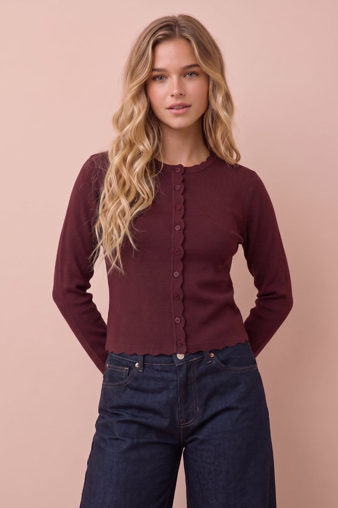 A woman with long wavy blonde hair wearing a dark burgundy button-up cardigan and dark blue jeans, standing against a solid dusty pink background.