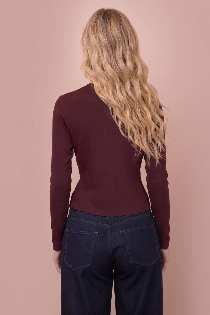 A woman with long blonde wavy hair is shown from behind, wearing a fitted long-sleeve ribbed top in deep burgundy and dark blue jeans, set against a solid dusty pink background.