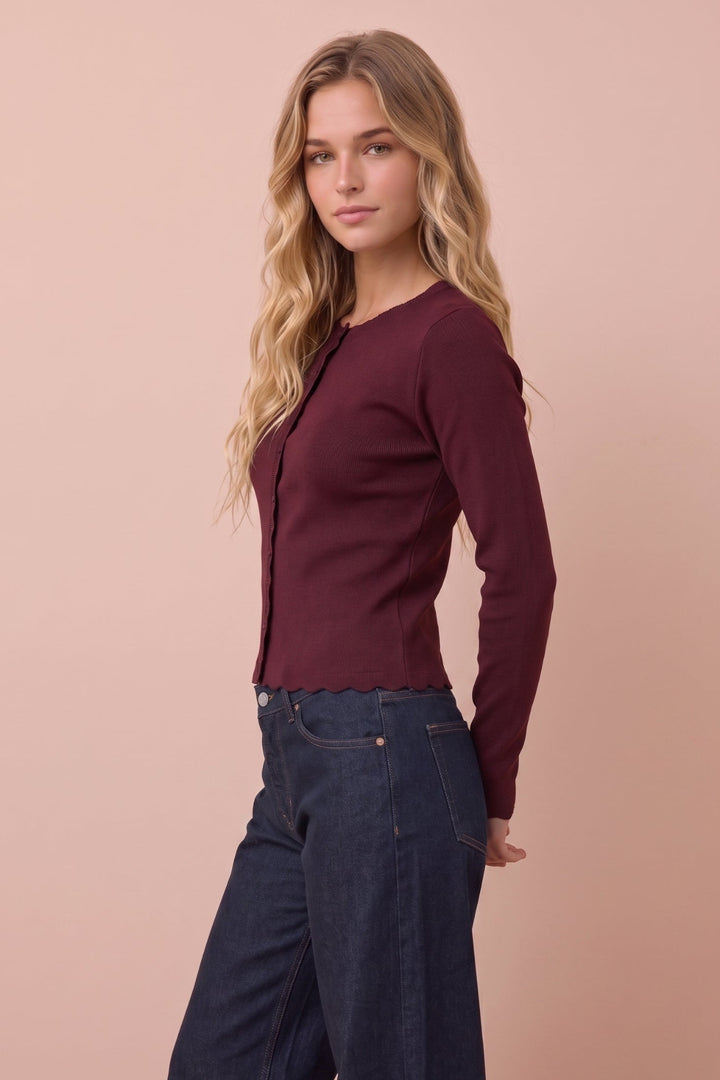 A woman modeling a long-sleeve ribbed knit top in deep burgundy, paired with dark blue wide-leg jeans, set against a solid pale peach studio background.