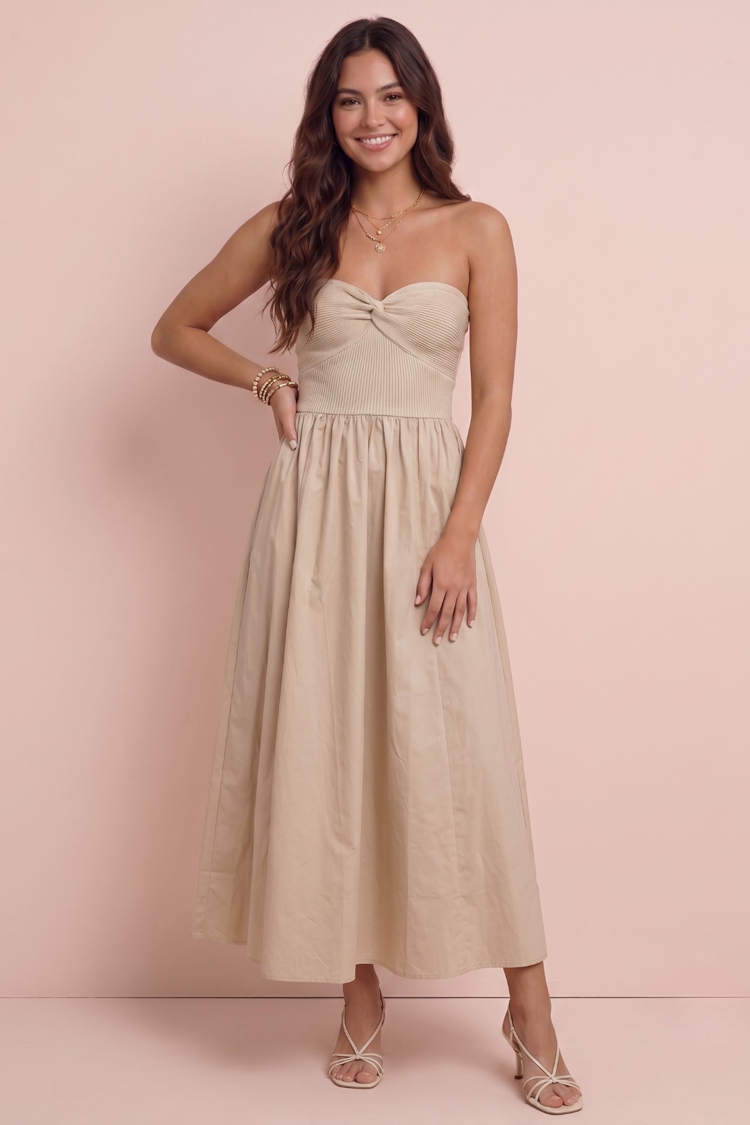 A woman modeling a strapless midi dress in a light beige or cream color, standing against a solid pale pink studio backdrop.